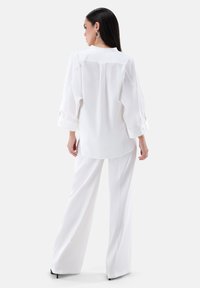 Weiße Langarmbluse mit weichem Fall, ausgestattet mit einem geknöpften Manschettenverschluss und geradem Schnitt. Kombiniert mit fließenden weißen Hosen.