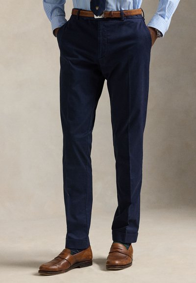 Man draagt marineblauwe broek, bruine leren riem, gestreept blauw hemd, marineblauwe das, horloge en bruine leren loafers, staat met handen in de zakken.