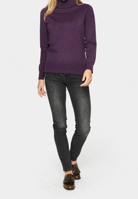 Femme portant un pull à col roulé violet, un jean skinny noir délavé et des mocassins en cuir marron à franges, debout devant un fond blanc.