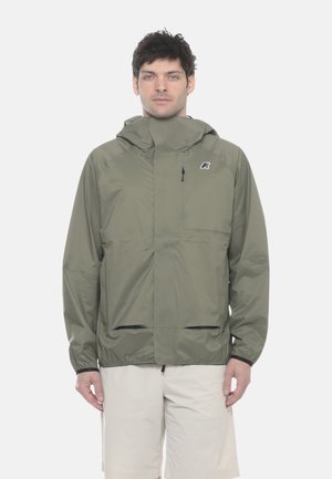 Mężczyzna w oliwkowozielonej kurtce przeciwdeszczowej z kapturem i beżowych szortach, stojący na jednolitym białym tle.