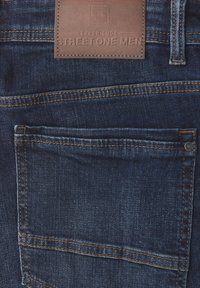 Tummansininen farkut sisältävät takataskun, jossa on erottuva ommel. Taskun ylle on kiinnitetty ruskea nahkaplakkari, jossa lukee "STREET ONE MEN".