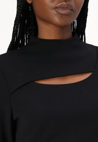 La robe noire présente un col haut avec un détail découpé au niveau de la poitrine. Le tissu semble lisse et texturé, avec des lignes épurées et une coupe ajustée.