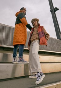 Zapatillas altas con una base blanca que presentan un estampado de leopardo negro y rosa. La modelo lleva pantalones de pierna ancha color crema y un suéter marrón.