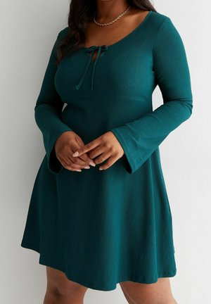 Femme portant une robe à manches longues couleur sarcelle avec des poignets évasés et un détail noué au niveau du col, debout devant un fond uni.