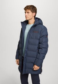 Giacca a vento blu navy con cappuccio, caratterizzata da sezioni trapuntate, un logo sul petto e tasche con zip. Texture liscia e vestibilità regolabile.