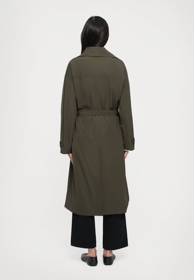 Olivengrüner Trenchcoat mit tailliertem Gürtel, langen Ärmeln und Schulterepauletten, kombiniert mit schwarzer, weiter Hose und flachen Schuhen.