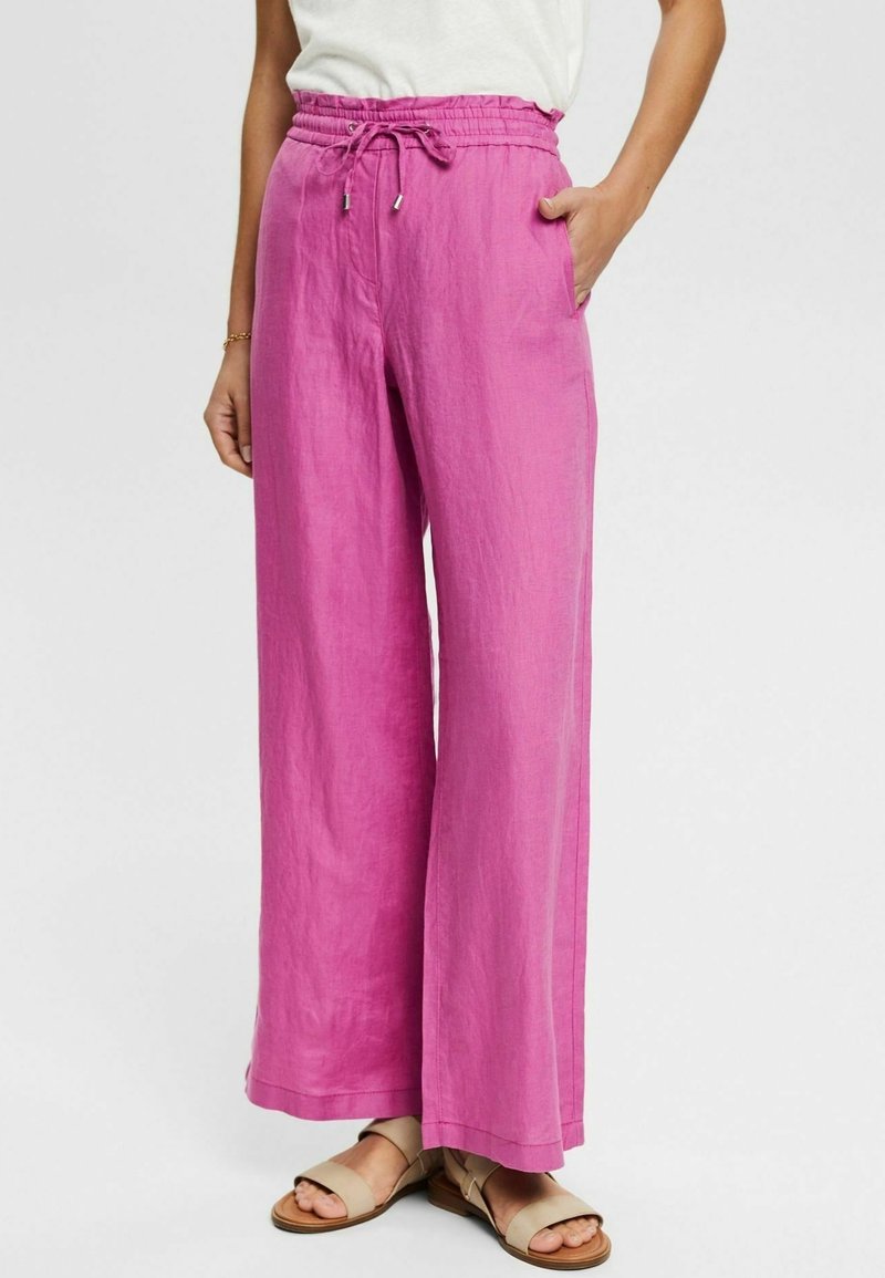 Femme portant un pantalon large en lin rose vif avec une taille élastique et un cordon de serrage, associé à des sandales beiges à bout ouvert.