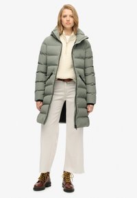 Lange, gewatteerde jas in gedempt groen, met een capuchon, ritsen en zijzakken. Draag het over een crèmekleurige gebreide trui en off-white wijde broek.