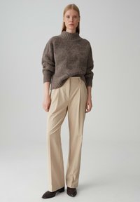 Bruine, oversized gebreide trui met hoge kraag gecombineerd met beige, geplooide broeken en donkere, puntige hakken. Soepele texturen, neutrale kleuren.