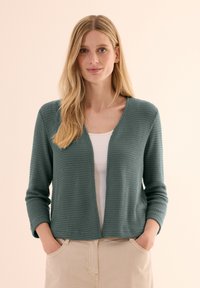 Vihreä teksturoitu neulecardigan, jossa on V-pääntie ja three-quarter hihat, päällä valkoinen toppi, yhdistettynä vaaleanruskeisiin housuihin.