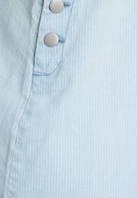 Prikaz blizu svetlo modre tkanine s subtilnimi vertikalnimi belimi črtami in dvema srebrnima pritisnima gumboma na šivu tkanine.