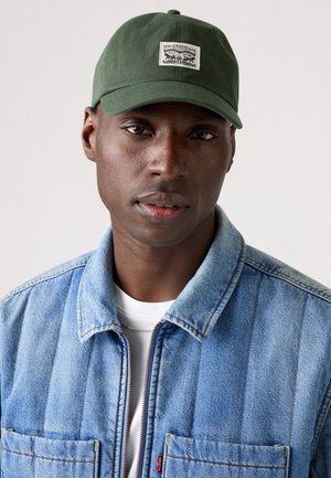 Green cotton baseball cap with a front label featuring a graphic design. Worn with a light blue denim jacket over a white shirt.
