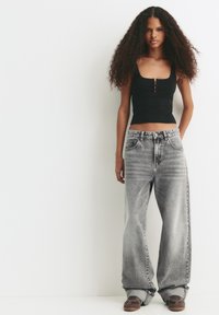 Jonge vrouw met lang, krullend haar, gekleed in een zwart mouwloos topje, oversized, vervaagde grijze jeans en een paar bruine sneakers, staand tegen een witte muur.