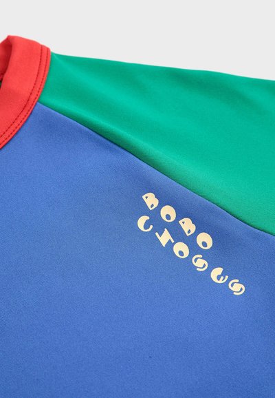 T-shirt multicolore con sezioni rosse, verdi e blu. Tessuto morbido con testo grafico di colore crema su fondo blu.