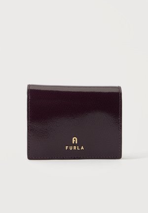 Mažas tamsiai violetinės spalvos lakinės odos piniginė su auksiniu Furla logotipu priekyje, šviesaus ir paprasto fono fone.