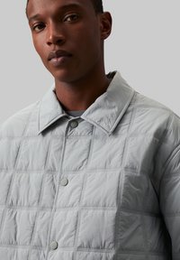 Hellgraue gesteppte Jacke mit Kragen, Knopfverschluss und einem strukturierten Stoff mit quadratischen Mustern.