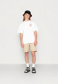 Balta pārizmēra t-krekls ar sarkanu grafiku, bēšas šortas, melna bļodiņas cepure, melnas un baltas sandales, baltas zeķes, stāv uz gluda fona.