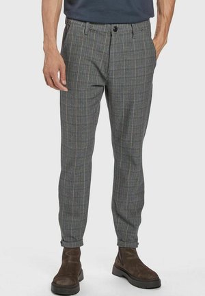 Homme portant un pantalon gris à motifs carreaux fuselé, associé à des bottes en daim marron, mains détendues le long du corps, sur un fond uni.