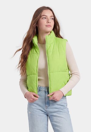 Jeune femme aux longs cheveux portant un gilet matelassé vert citron sur un pull côtelé beige et un jean bleu clair, regardant légèrement sur le côté.