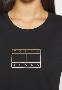 Černé tričko s zlatým obdélníkovým logem, na kterém je text "TOMMY" nad "JEANS." Tričko má kulatý výstřih a krátké rukávy.