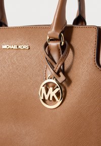 Καφέ δερμάτινη τσάντα χειρός με υφή επιφάνειας, χρυσά μεταλλικά στοιχεία και στρογγυλό έμβλημα λογότυπου. Το όνομα της μάρκας είναι ανάγλυφο σε χρυσό χρώμα.