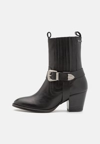Bota de cuero negro hasta el tobillo con punta afilada, costuras elevadas y un acento de hebilla. Tacón bloque y lazos de tirón para facilitar la puesta.