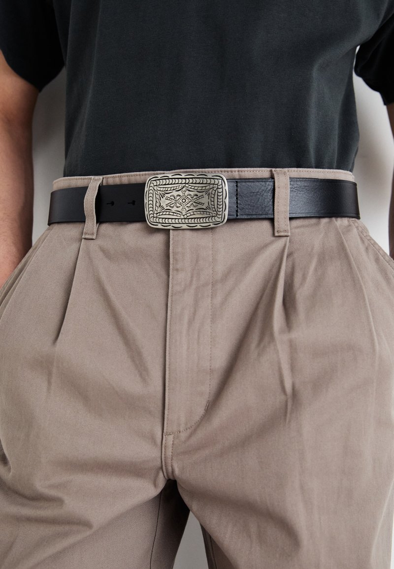 Ceinture en cuir noir ornée d'une boucle en argent élaborée avec des motifs complexes, associée à un pantalon plissé beige clair.
