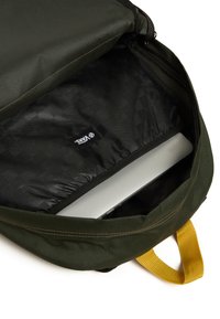 Interieur van een groene stoffen rugzak, met een zwart gevoerd vak waarin een grijze laptop zichtbaar is, en een geel draaghandvat.