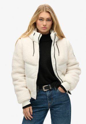 Witte en crême gevoerde jas met een donzige textuur, hoge kraag en zwarte ritsen, gecombineerd met een donkere trui en blauwe jeans.
