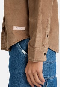 Braune Cordhemd mit senkrechten Rippungen, mit Knopfmanschetten und einem Etikett mit der Aufschrift "ELEMENT CO. SINCE 1992" in der Nähe des Saums. Getragen über blauer Jeans.