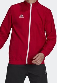 Rød letvægts atletisk jakke med opretstående krave, hvid lynlås og en diskret tre-stripes logo på den øverste bryst. Teksturen virker glat.