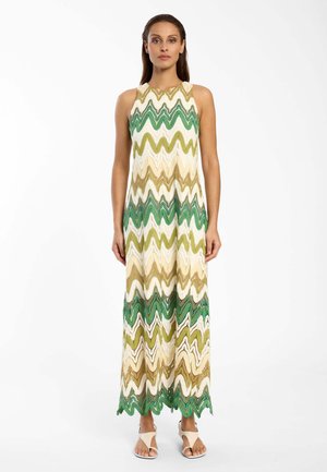Vrouw draagt een mouwloze jurk tot op de grond met een zigzagpatroon in groen, beige en wit, staand met ontspannen armen langs haar zij.