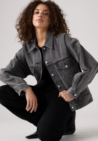Gray denim jacket with a relaxed fit, two front pockets, silver hardware, and a slightly faded look, paired with black pants.