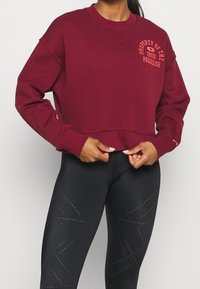 Felpa corta bordeaux con polsini a costine e scollatura a girocollo, caratterizzata dalla stampa "Proprietà dell'Iron Paradise" in arancione. Abbinata a leggings neri decorati con un motivo perforato geometrico.