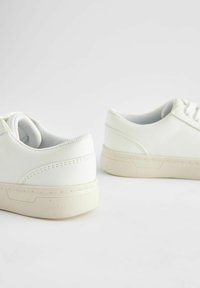 Bílé kožené tenisky s nízkým profilem, texturovanými akcenty a polopropustnou gumovou podrážkou. Obsahují tradiční šněrovací design.