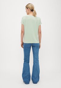 Lys grøn t-skjorte med korte ærmer laget av glatt stoff, kombinert med blå jeans med vid bunn. Designet har rullede ærmer og en avrundet baktetning.