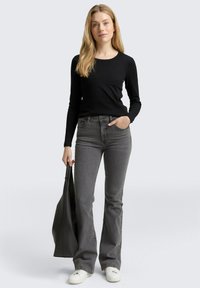 Schwarzes, langärmeliges, geripptes Oberteil, graue Schlagjeans und weiße Sneakers. Das Modell hält eine große schwarze Umhängetasche und präsentiert einen lässigen Look.