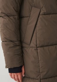 Brun pufferjakke med quiltet design, der har en sidelomme og ribstrikkede manchetter. Materialet ser glat og isoleret ud for varme.
