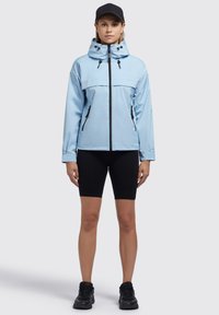 Veste imperméable bleu clair avec un col haut, fermeture éclair noire et accents, associée à un short de vélo noir et des baskets noires.