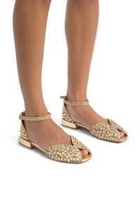 Sandali flat dorati con una fascia trasparente, decorati con gruppi di pietre strass, dotati di punta aperta e tacco quadrato.