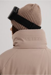 Beige jakke med høj krave og subtilt prægede logo, parret med en ribbet beige strikket hue og et sort gogglesnor.