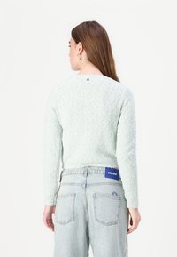 Light green, textured sweater with long sleeves, round neckline, and a small logo detail at the back, paired with high-waisted light blue jeans.