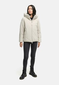 Donna che indossa un giubbotto imbottito beige chiaro con colletto alto, jeans skinny neri e stivali neri con lacci, in piedi davanti a uno sfondo bianco.