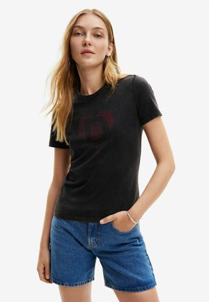 Camiseta de algodón negra con mangas cortas, que presenta un diseño de corazón rojo con una letra en el interior. Combinada con pantalones cortos de mezclilla azules.