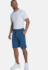Blaue Baumwollshorts mit elastischem Bund und Kordelzug, ausgestattet mit Seitentaschen und einem Logo-Patch. Kombiniert mit einem figurbetonten weißen T-Shirt und Turnschuhen.