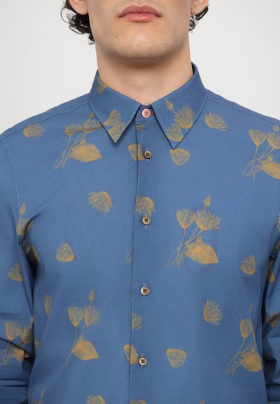 Camisa azul de botones con un patrón floral dorado, cuello puntiagudo y botones rosas. Hecha de una tela suave con un corte regular.