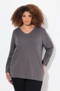 Donna con capelli ricci che indossa una maglia grigia larga a maniche lunghe con scollo a V e pantaloni neri, in piedi davanti a uno sfondo bianco.