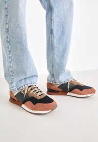 Zapatillas con una mezcla de tela negra, ante marrón y acentos color crema. Presentan una suela texturizada y cierre de cordones. Se llevan con jeans de color azul claro.