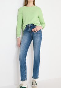 Femme portant un pull vert clair, un jean bleu, des chaussettes blanches et des baskets beige avec des détails verts, debout contre un mur blanc.