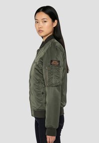 Olivengroene bomberjack met een gladde, glanzende textuur, geribbelde manchetten, en een ritszak aan de linkermouw met een patch.